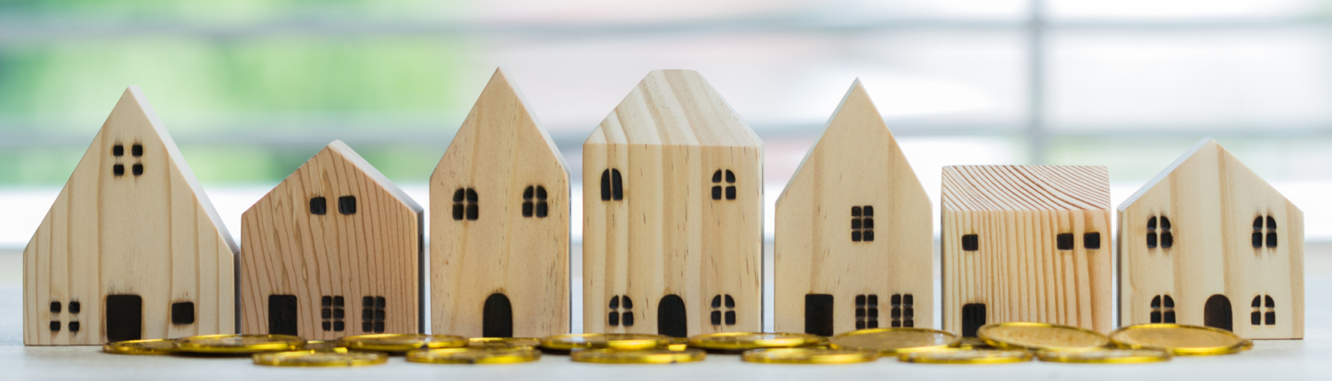 Finanzierung. Eine Reihe von Holzmodellhäusern mit Goldmünzen im Vordergrund - Martina Wagner Immobilien