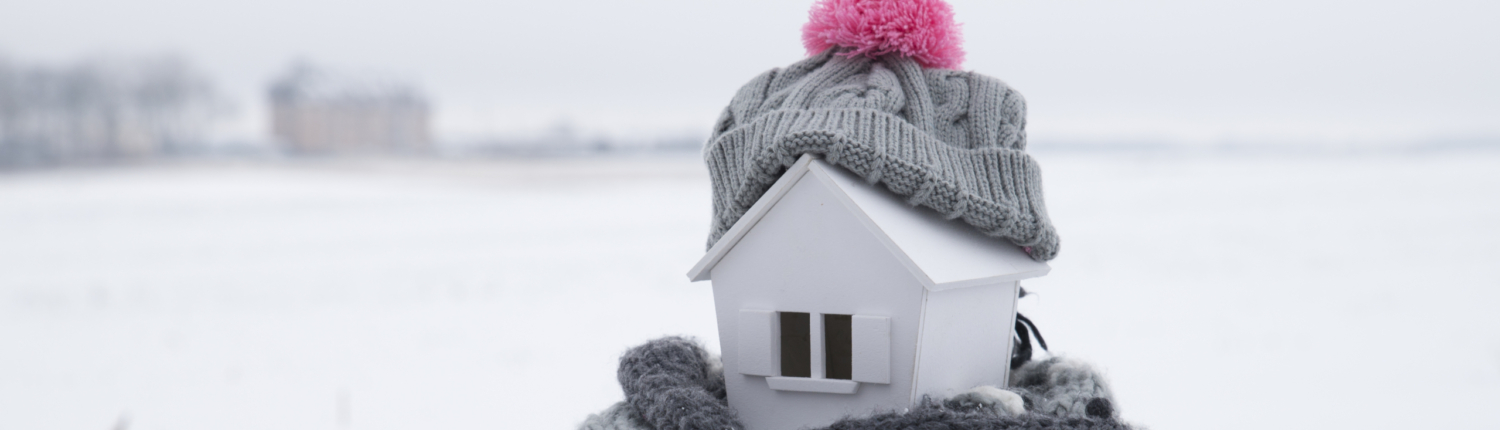 Ein Hausmodell mit einer Mütze auf dem Dach und einen Schal am Boden steht im Schnee - Martina Wagner Immobilien