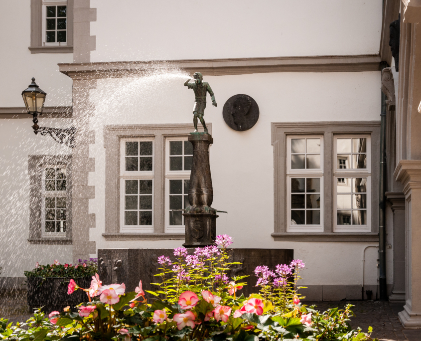 Schängel Brunnen in der Altstadt von Koblenz bei Sommerwetter mit blühenden Blumen im Vordergrund - Martina Wagner Immobilien