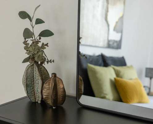 Teile eines modern eingerichteten Schlafzimmers werden durch einen Spiegel gezeigt, der auf einem schwarzen Tisch steht, daneben sind Pflanzen in Vasen dekoriert - Martina Wagner Immobilien