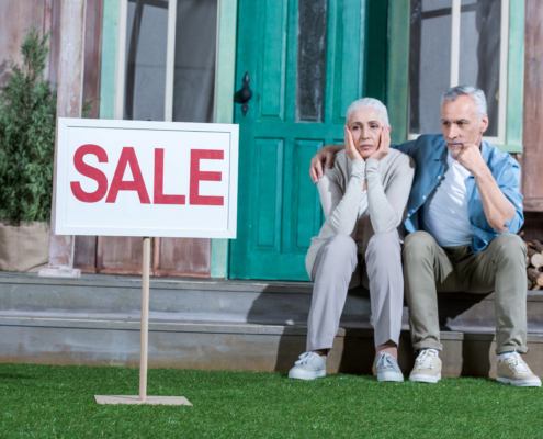 älteres Ehepaar sitzt auf der Treppe vor einem Haus, links neben steht ein "zu Verkaufen Schild".