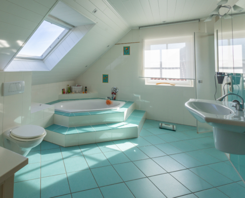 großes Bad mit 2 Fenster, türkisfarbenen Bodenfliesen und hellen Einbauten, eine zentrale Eckbadewanne, die Toilette und ein Waschbecken sind zu sehen.