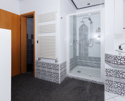 modernes Bad, rechts Waschbecken im Anschnitt mit besonderen schwarz-weiß Fliesen, daneben links Dusche, daneben Handtuchwärmer, daneben Zugangstür zum WC