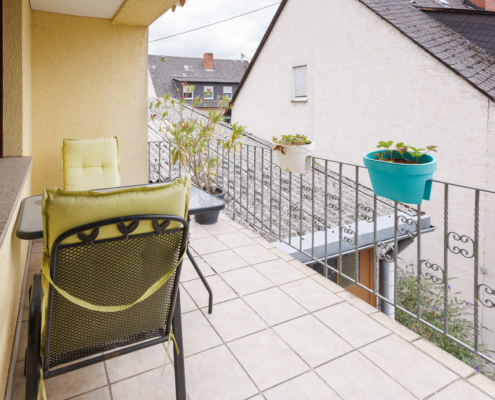 Balkon, Fenster links, davor kleiner Tisch mit 2 Stühlen mit grünen Bezügen, Boden ist gefliest, Geländer Gusseisen mit einigen Pflanzen. Man blickt auf das Nachbargebäude