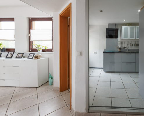 Blick durch ein Glasfenster auf eine moderne graue Küchenzeile, Zugang zur Küche links daneben, links daneben 2 braune Fenster, darunter weiße Kommode
