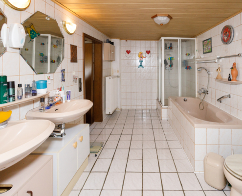 innen liegendes, hell gefliestes Bad mit Wanne, Dusche und Toilette auf der rechten und Doppelwaschbecken mit Spiegeln auf der linken Seite
