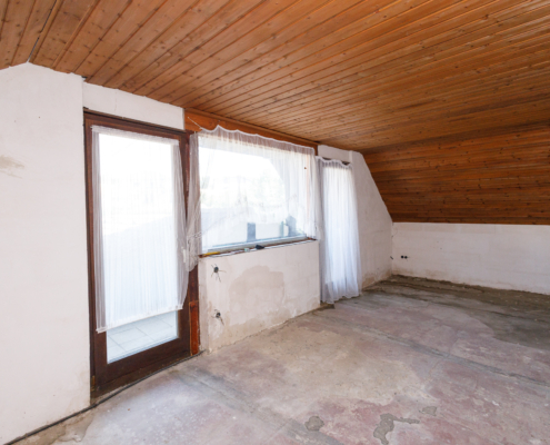 Rohbauansicht des Wohnzimmers, man sieht die Holzpaneeldecke und den Zugang zum Balkon über 2 Glastüren mit Fenster in der Mitte.