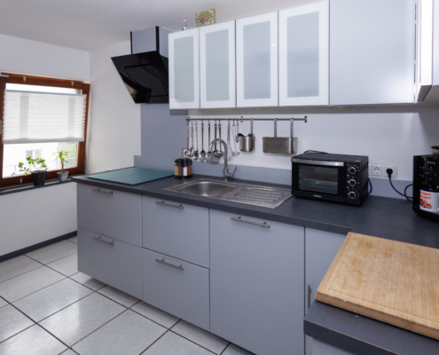 graue zeitlose Küchenzeile mit schwarzer Arbeitsplatte, links braunes Fenster, Fliesenboden hellgrau, rechts liegt ein Holzbrett auf der Arbeitsfläche, dahinter Kaffeemaschine und Mikrowelle