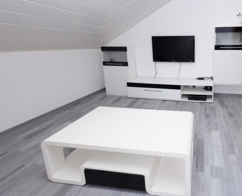 modernes Zimmer, grauer Laminatboden, Wände weiß, Dachschräge links mit weißen Holzpaneelen, im Vordergrund moderner weißer Couchtisch dahinter weiße TV Wand mit schwarzen Applikationen