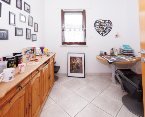 kleines Zimmer, helle Fliesen, weiße Wände, kleines Fenster, rechts braune Tür, davor kleiner Tisch und Drehstuhl, rechts braune Kommode mit vielen Bildern