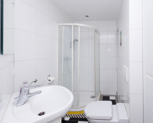 Duschbad, Kariertes Fliesenmuster in schwarz-weiß, ansonsten ist alles weiß, deckenhoch gefliest, links vorne Waschbecken, rechts Toilette, hinten Runddusche mit gelbem Sockel.