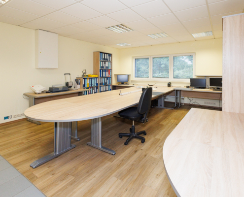 Büroraum mit großem vorne abgerundeten Schreibtisch mittig, rechts kleinerer abgerundeter Schreibtisch, im Hintergrund, Regale und Schränke sowie Fenster