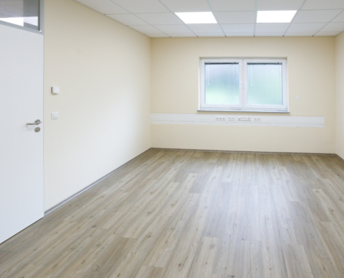 Büro mit Fenster im Hintergrund, darunter IT Leiste, Boden helles Holz, Links weiße Tür