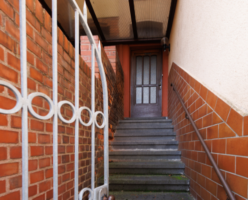 Man sieht einen Treppenaufgang zu einer Haustür, die rechte Seite ist unten braun gefliest, darüber verputzt, links erkennt man eine Ziegelmauer und ein Eisentor.