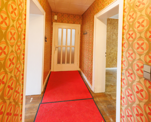 Man sieht einen Flur mit 70er Jahre Tapete, einem roten Teppich auf dem Boden. Rechts und links jeweils eine offene Tür und im Hintergrund eine geschlossene Tür mit Glaseinsätzen.