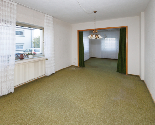 Wohn-Esszimmer mit 2 großen Fenstern links, 1 Fenster geradeaus. Es liegt ein grüner Teppich und am Durchgang befindet sich ein grüner Vorhang.