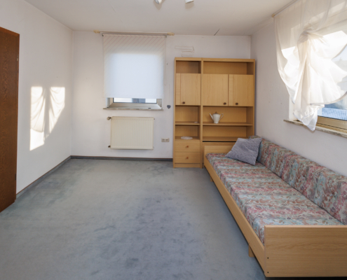 Zimmer mit 2 Fenstern, blauem Teppich, rechts ist ein Sofa, dahinter steht ein Schrank. Links ist eine Tür zu sehen.