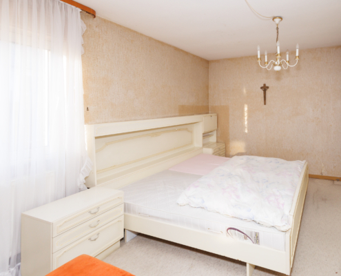 Blick in ein Schlafzimmer mit großem hellen Bett und Nachtschränken sowie großem Fenster mit weißen Vorhängen links. Rechts ist der Kleiderschrank zu erkennen.