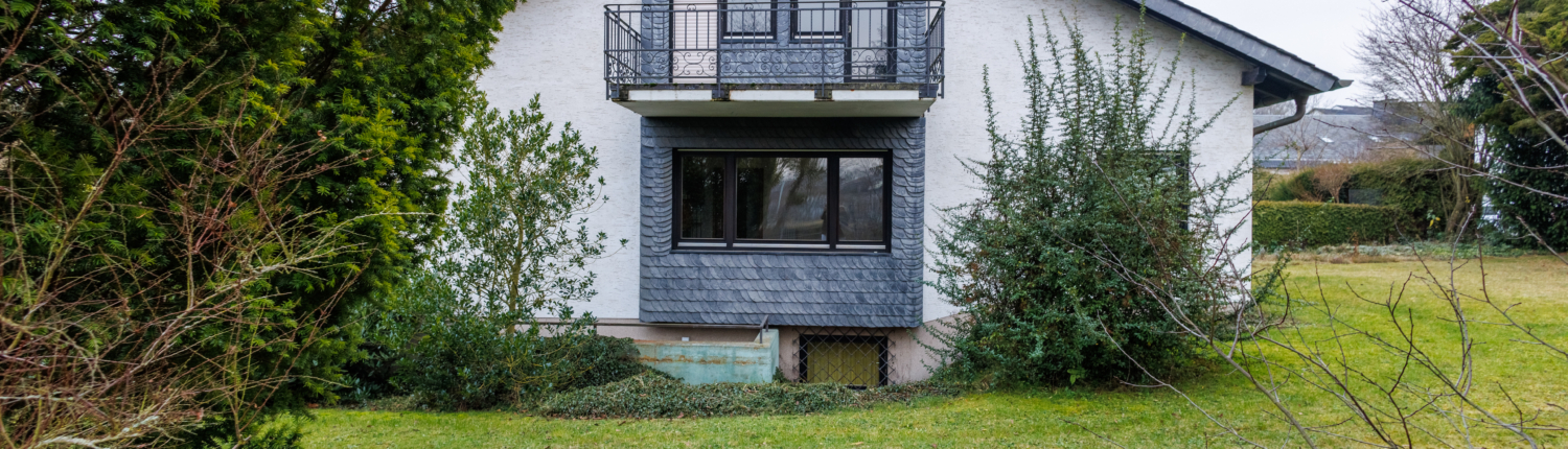 Seitenansicht Haus mit geschiefertem Bereich und Balkon, davon Büsche, Wiese